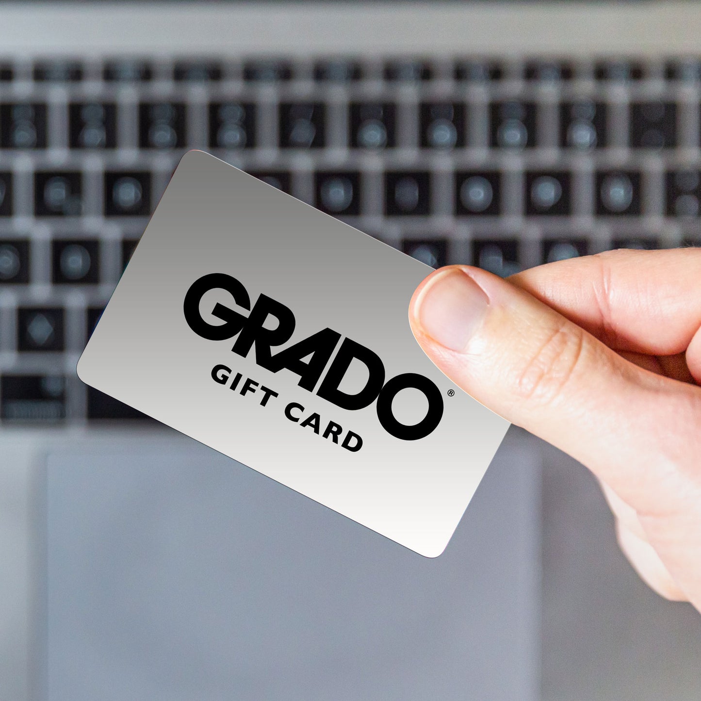 Photo of a a person holding a silver grado gift card with black letters on it that say "GRADO GIFT CARD". The background has blurred black boxes on it.