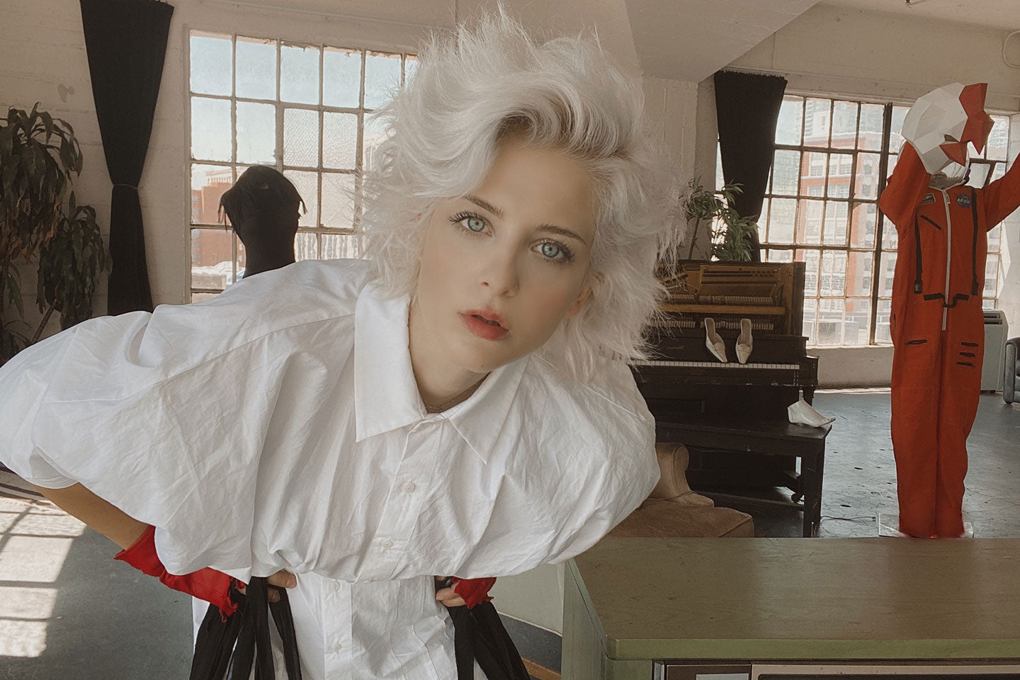 Photo of  Buel with white hair and a white shirt leaning over and staring at the camera. there are large windows and a city skyline in the background.