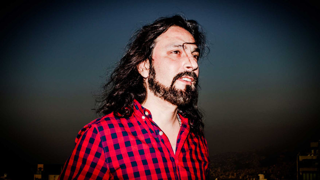 Photo of a man with a black beard and long hair. He is wearing a black and red checkered shirt and standing in front of a twighlight sunset.